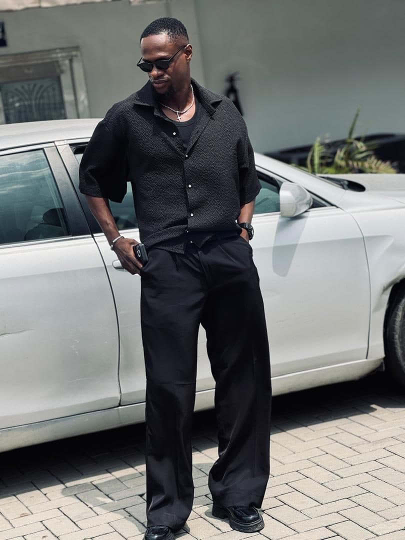 Black shirt and wide black trousers, worn with sunglasses beside a white car